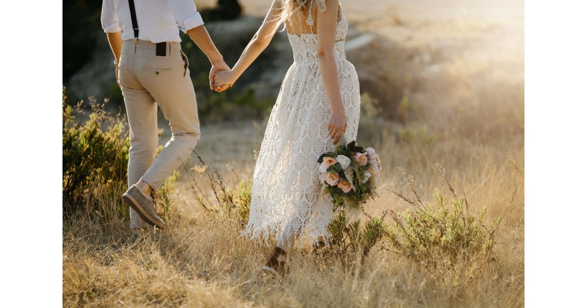 A couple holding hands while walking through a field, the woman in a lace dress holding a bouquet of pink roses, with the sun shining in the background.