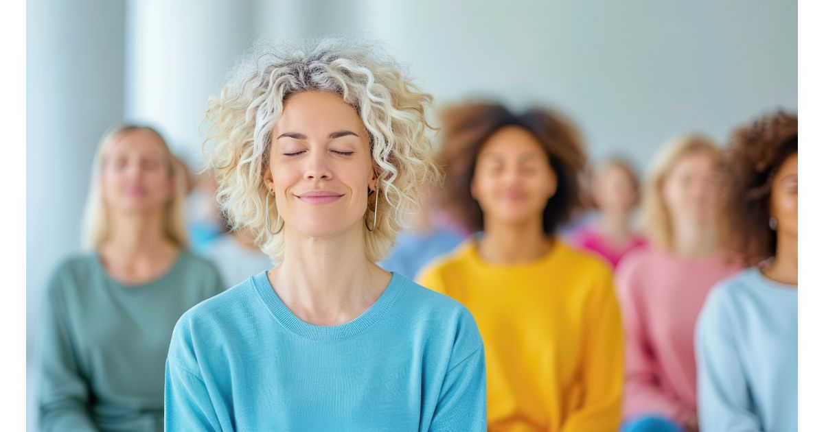 A woman with curly hair calmly meditating in a group, with others in the background also participating in a relaxation or mindfulness session.