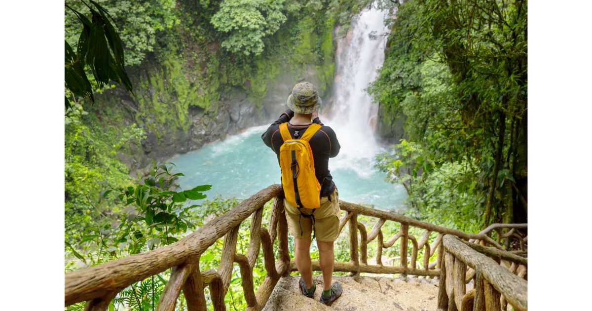 A person standing at a wooden railing, taking photos of a beautiful waterfall surrounded by lush greenery. The vibrant blue water and tropical forest create a stunning scene.