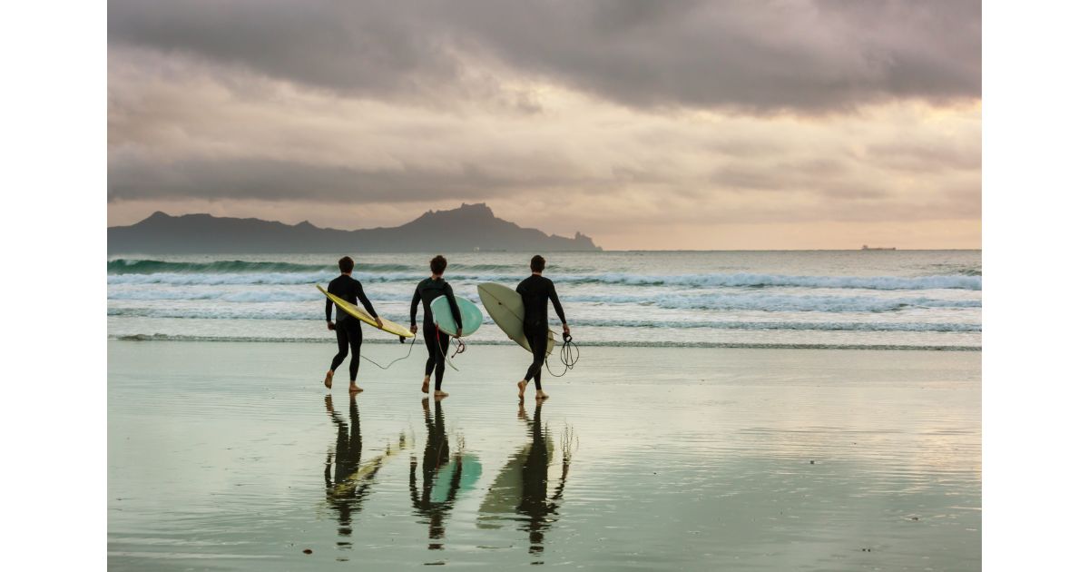 Three surfers walking along the beach with their surfboards, the cloudy sky reflecting on the wet sand. The waves are calm, and the silhouette of a distant island can be seen.