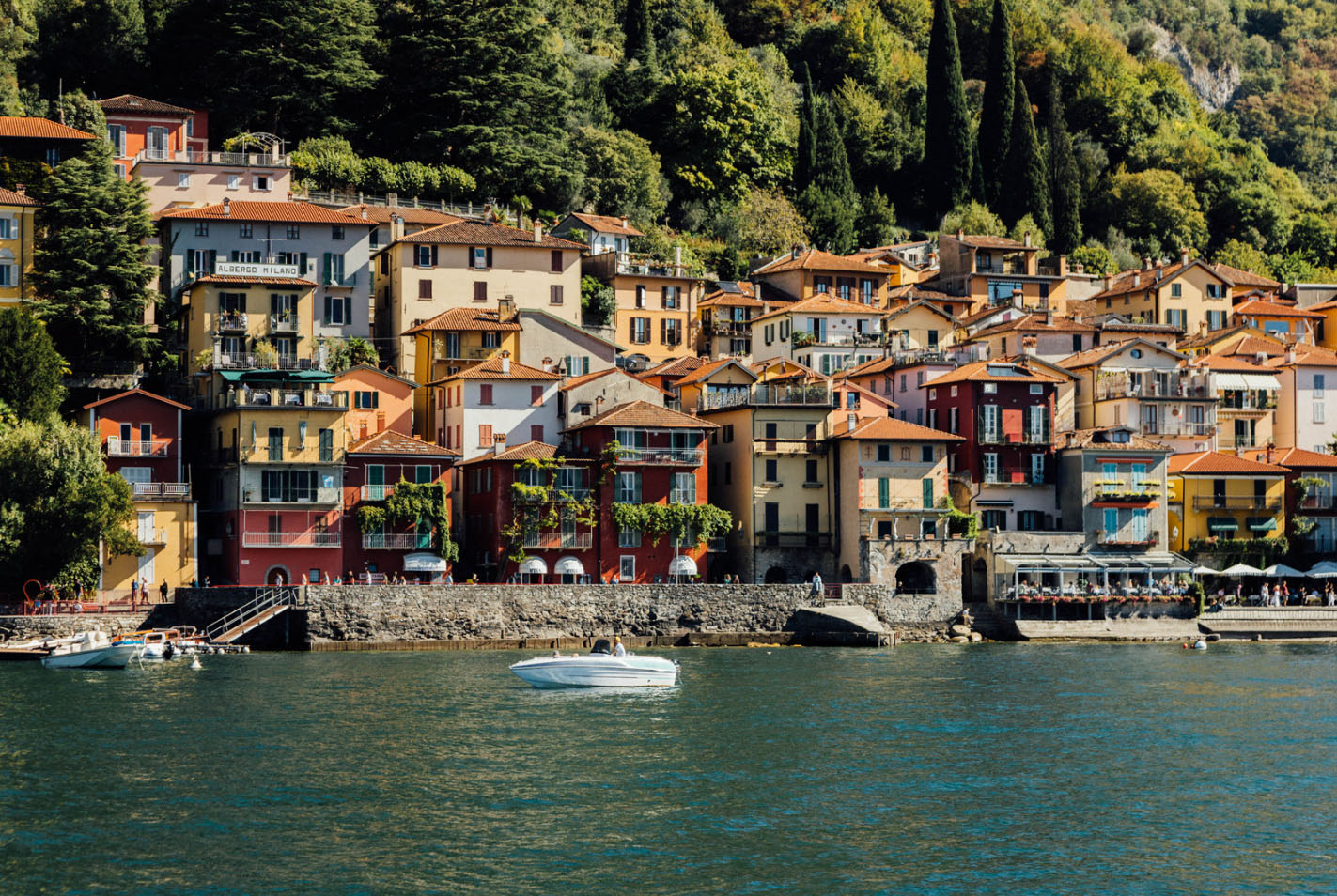 A scenic view of colorful lakeside villas in Lake Como, Italy, with a white boat cruising on the calm waters, surrounded by lush greenery and mountains.