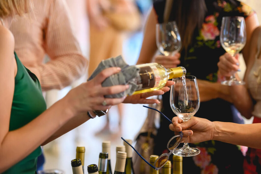 A close-up of a wine tasting event, where a server is pouring white wine into a glass held by a guest, surrounded by people holding wine glasses.