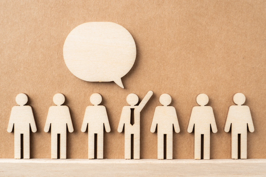 Wooden figurines of people with one figure raising its hand inside a speech bubble, representing communication or speaking up.