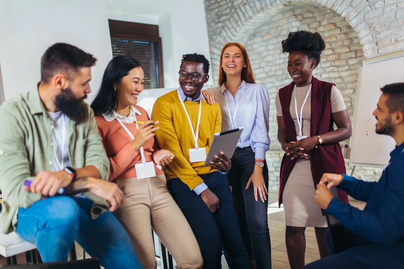 A diverse group of professionals smiling and interacting during a team-building or business event, with one person holding a tablet and others engaged in conversation.