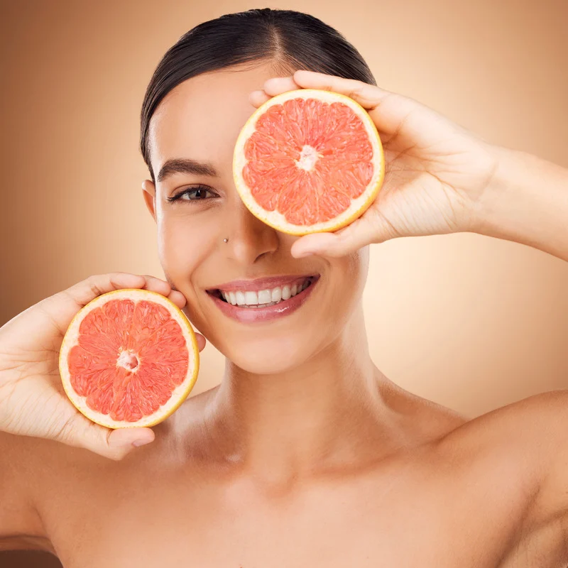 A smiling woman holding two halves of a grapefruit in front of her face, symbolizing health and wellness. She has radiant skin and a glowing smile.