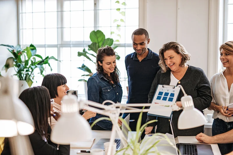 A group of diverse professionals collaborating in a bright office space, with one person holding a presentation board and others engaged in discussion around a table.