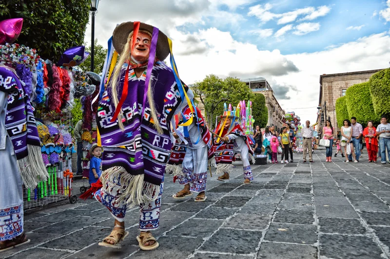 People wearing traditional Mexican costumes and large colorful masks dance through a street during a cultural celebration, with onlookers and festive decorations in the background.