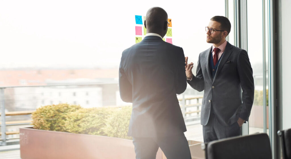 Two business professionals in formal suits engaged in a discussion near a window with a cityscape view. Colorful sticky notes are arranged on the glass.