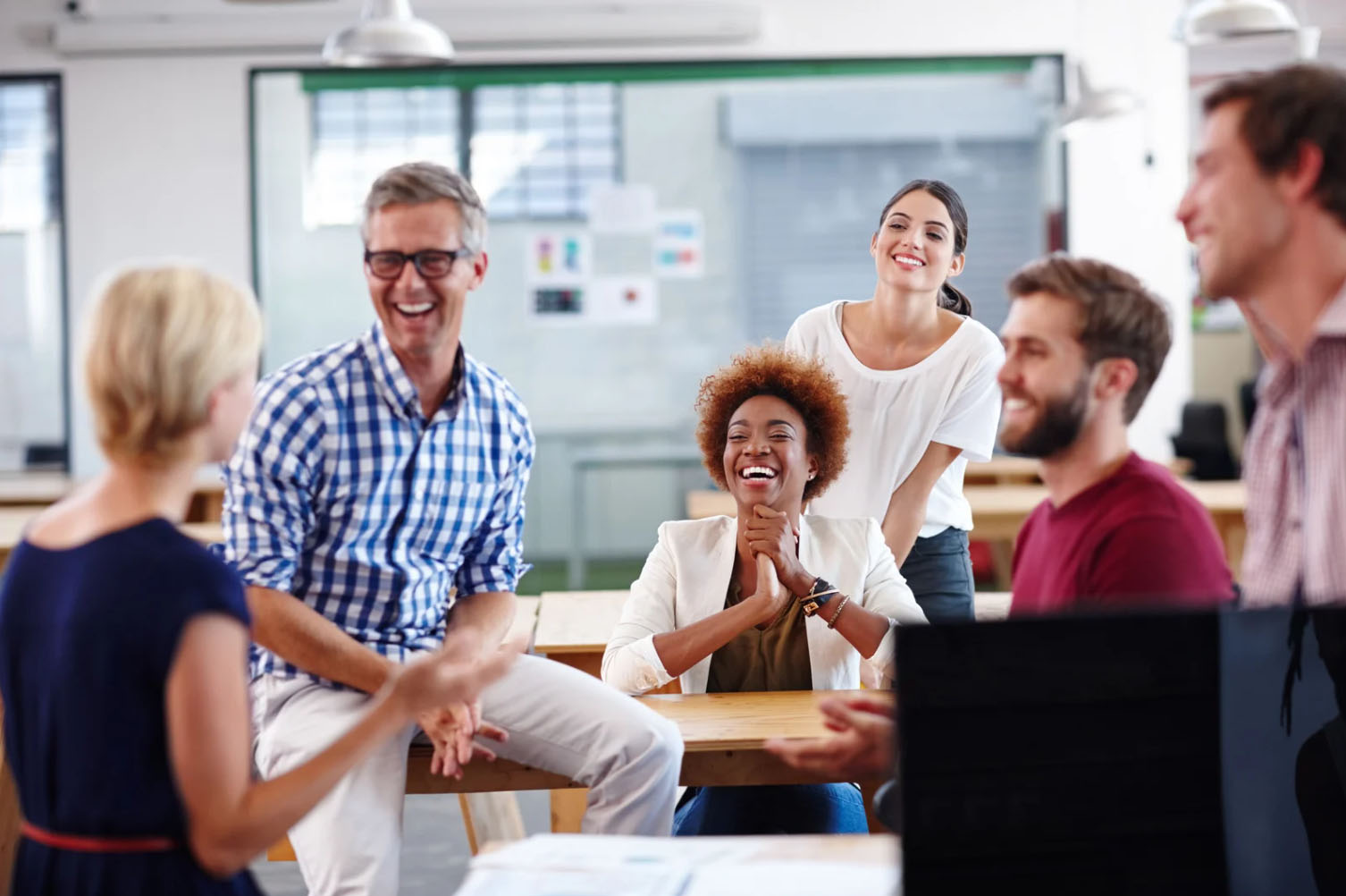 A diverse group of professionals laughing and engaging in a discussion in a modern office setting, showcasing a positive and energetic workplace environment.