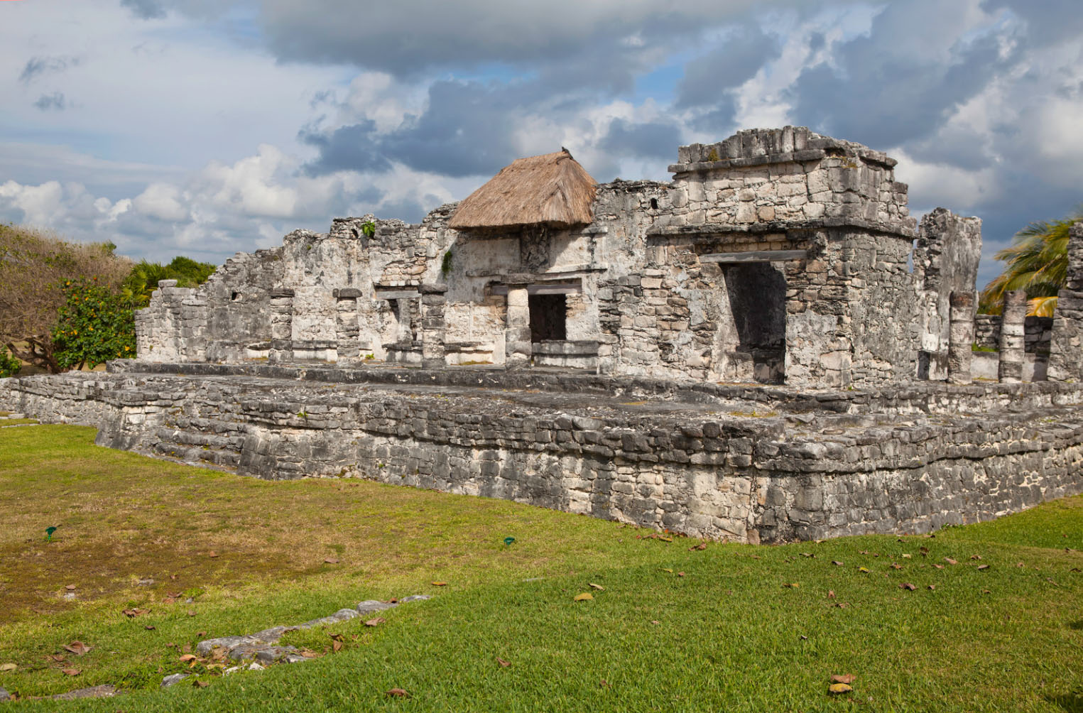Ancient stone ruins of a Mayan temple in Tulum, Mexico, featuring a thatched roof structure, surrounded by lush green grass under a cloudy sky.