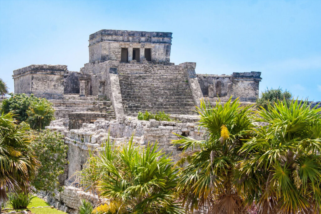 Ancient Mayan ruins of Tulum with stone structures and stairways surrounded by lush green palm trees under a bright blue sky.