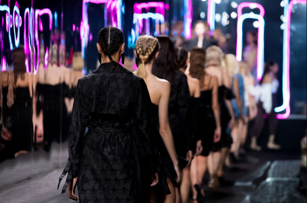 Models walking down the runway at Milan Fashion Week, showcasing high-end fashion against a backdrop of neon lights and a stylish audience.
