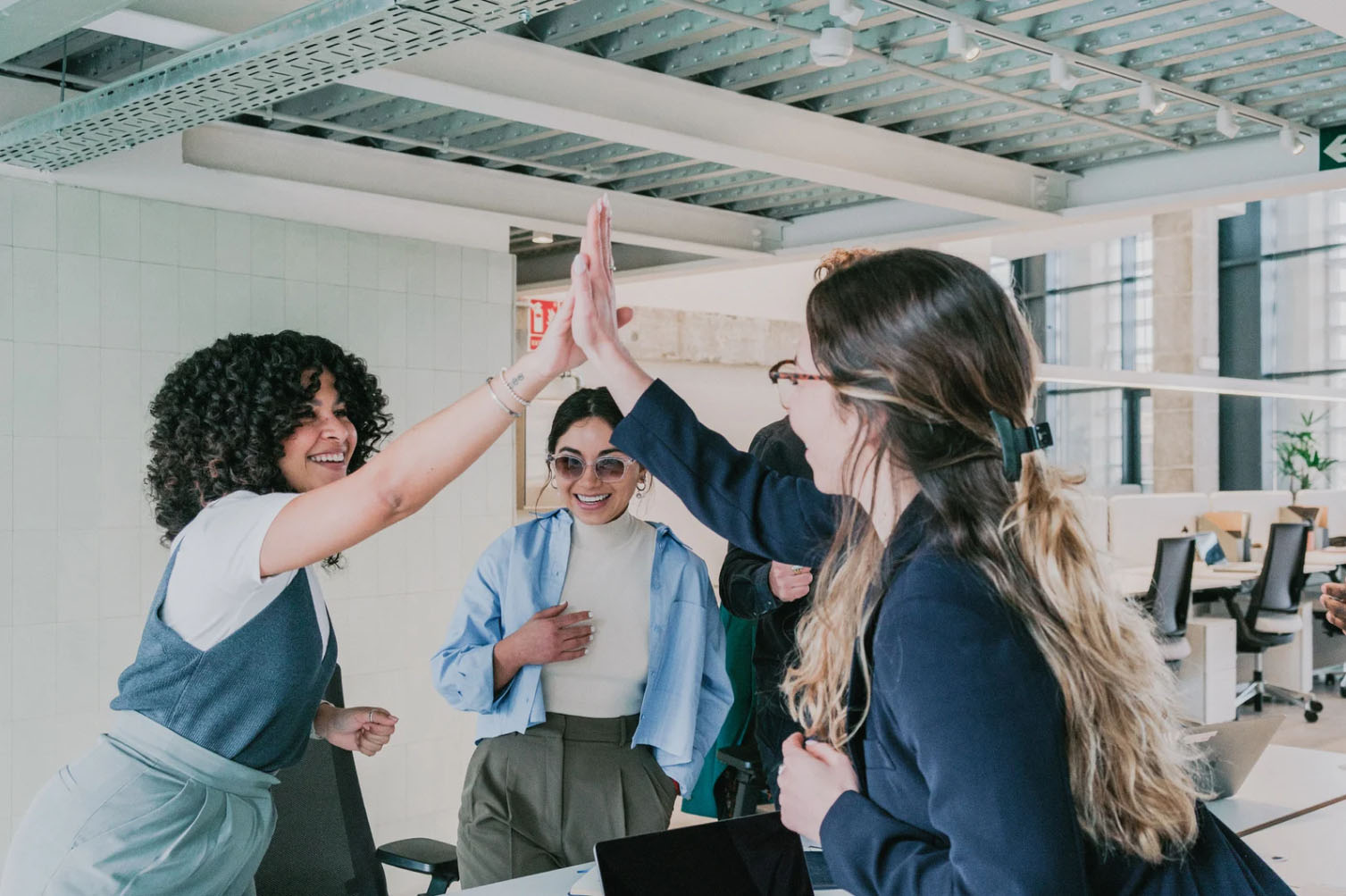A group of diverse professionals in an open office space celebrating teamwork with a high-five, smiling and engaging in a positive work environment.