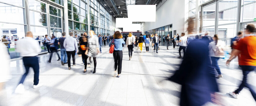 A blurred image of a crowded convention center or trade show with attendees walking in various directions.