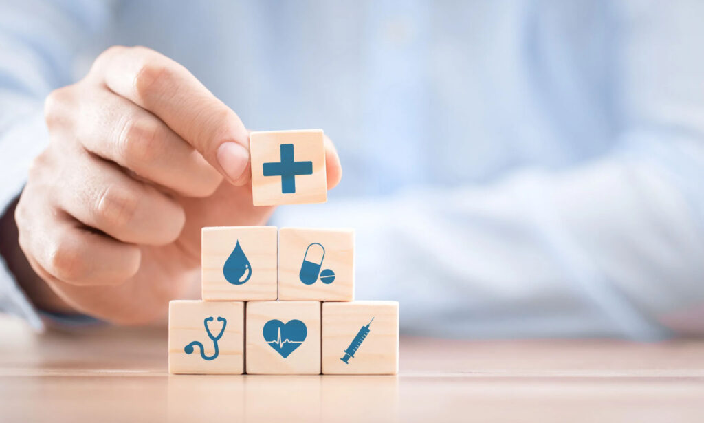 A hand placing a medical cross block on top of stacked blocks featuring health-related icons, including a stethoscope, heart, syringe, water droplet, and pills.