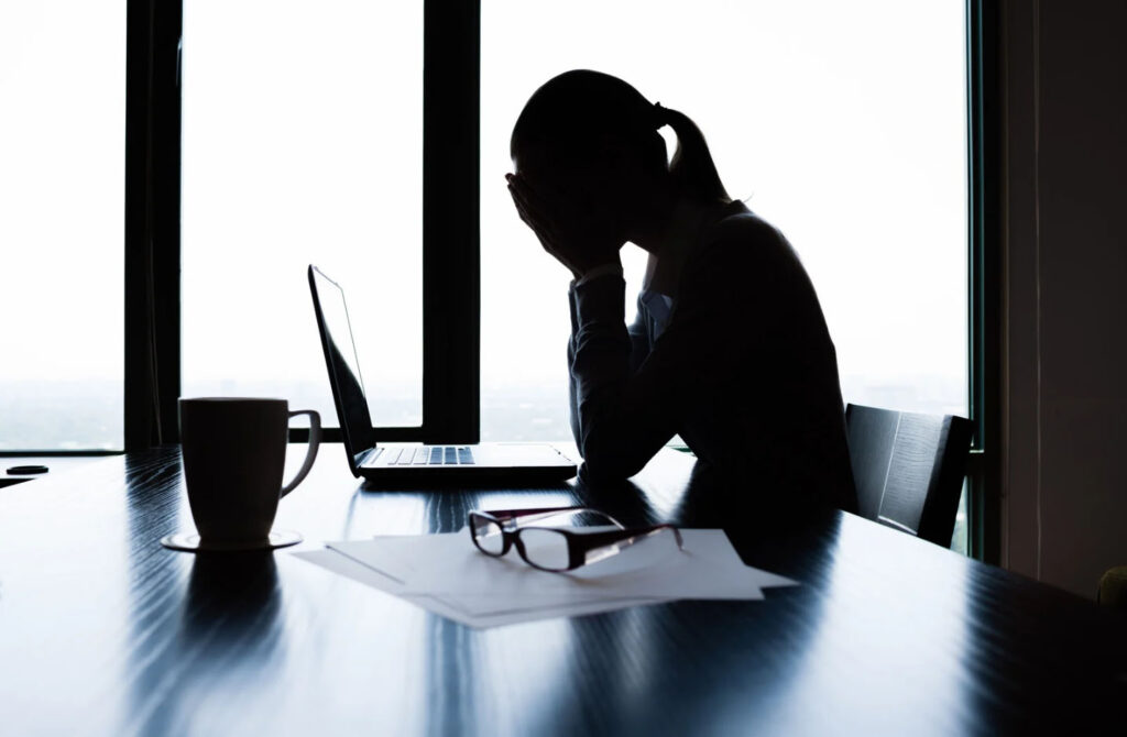 Silhouetted woman sitting at a desk with her head in her hands, appearing stressed or overwhelmed, with a laptop, papers, and glasses on the table.