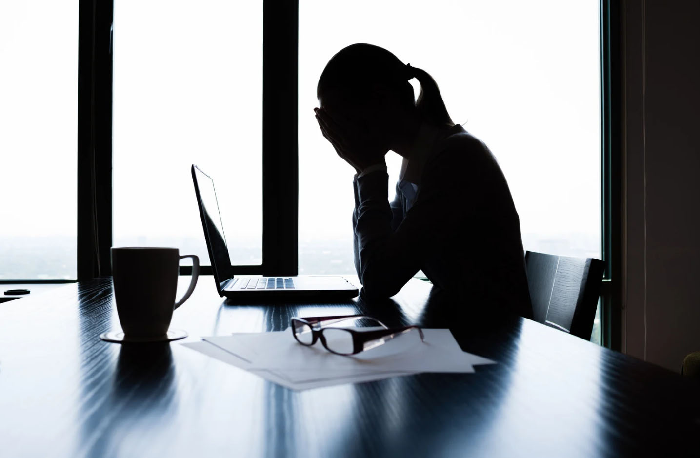Silhouetted woman sitting at a desk with her head in her hands, appearing stressed or overwhelmed, with a laptop, papers, and glasses on the table.