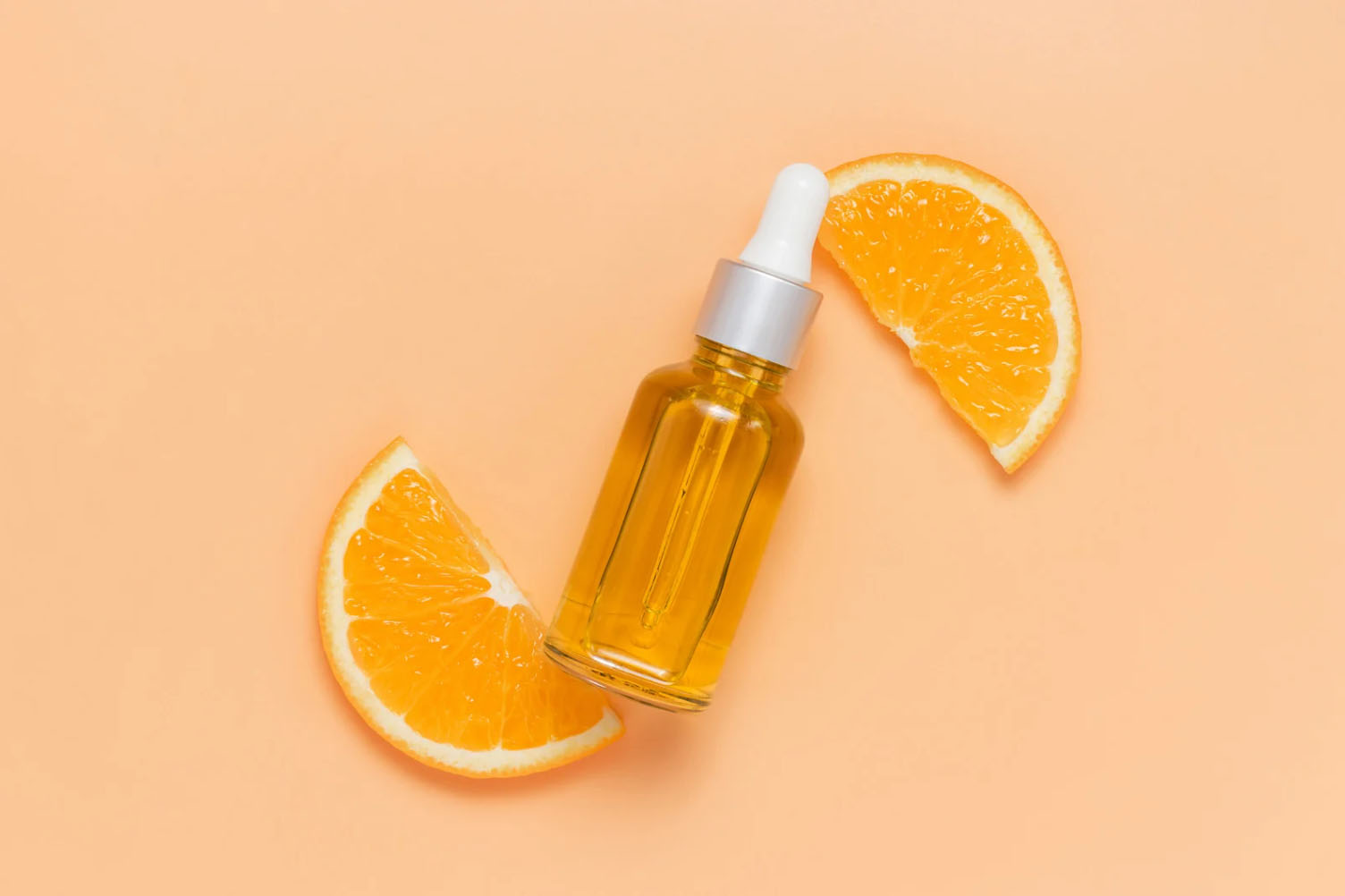 Dropper bottle of essential oil with fresh orange slices on a peach-colored background.