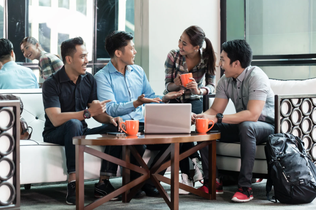 Four people in casual attire engaging in a conversation with coffee cups while sitting around a table with a laptop in a modern office setting.