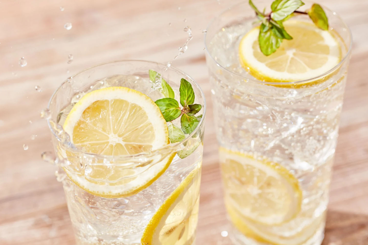 Two glasses of sparkling water with lemon slices and fresh mint, with water splashing into one glass on a wooden surface.
