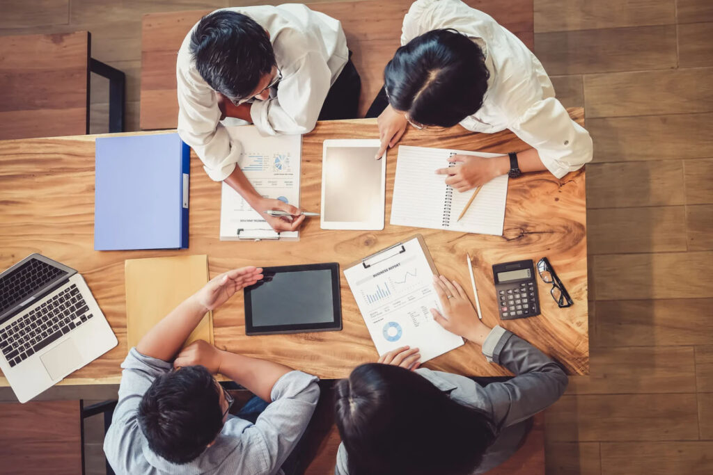 Overhead view of four people sitting around a wooden table, collaborating with documents, tablets, a notebook, and a calculator.