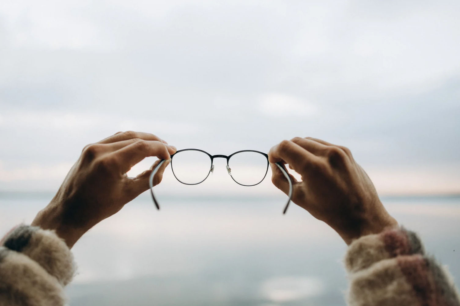 Person holding eyeglasses with both hands against a blurred background of a calm body of water and cloudy sky.