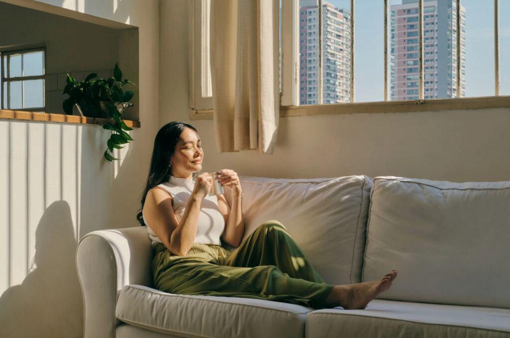 Woman sitting on a white couch in a sunlit room, eyes closed and smiling while holding a cup, with tall buildings visible through the window.