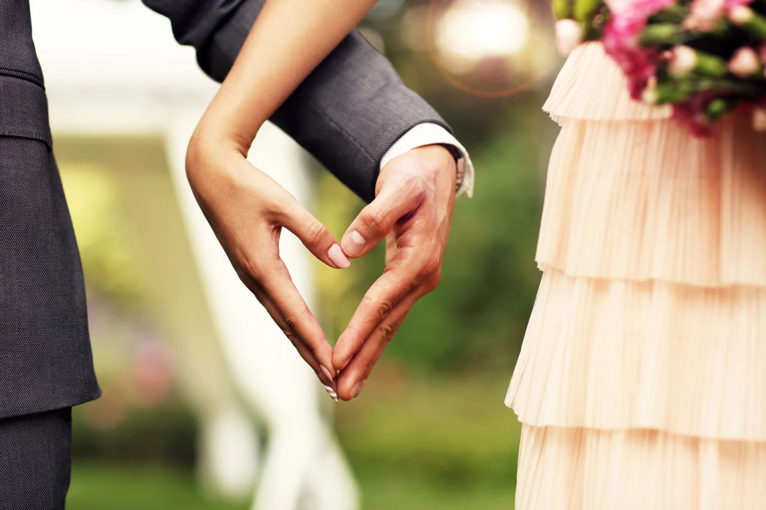 A close-up image of a bride and groom holding hands, forming a heart shape with their fingers, with a soft, blurred background.