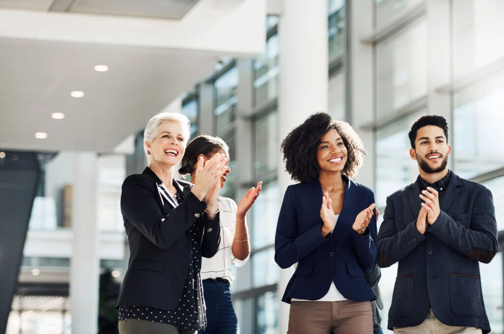 Four diverse professionals, two women and two men, clapping together in a bright, modern office setting.