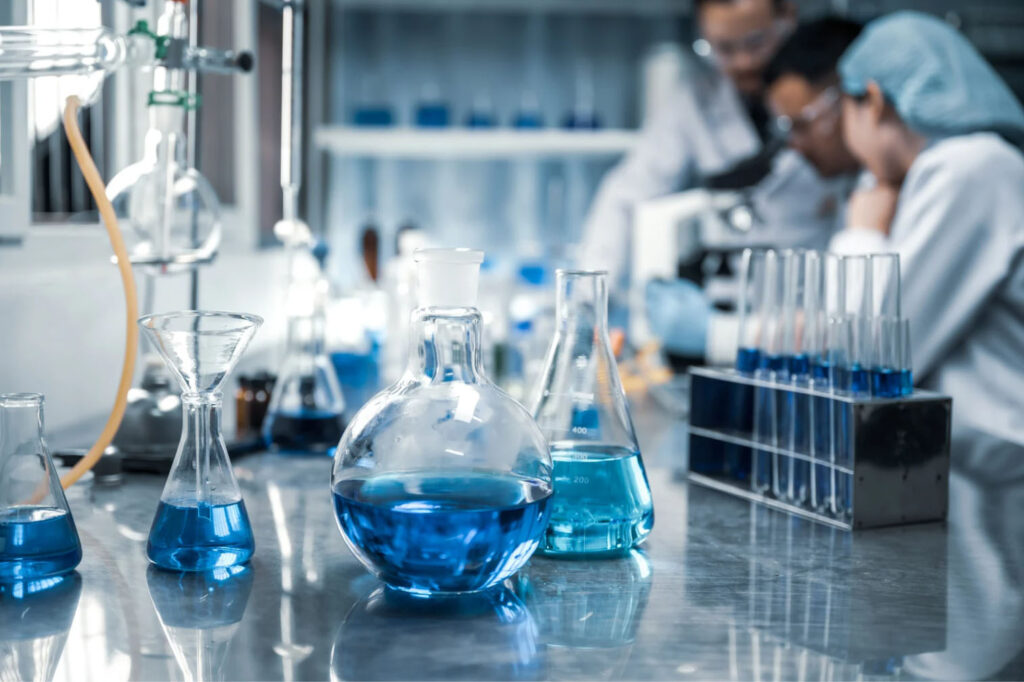 Laboratory setting with scientists working in the background. Several glass beakers and flasks containing blue liquid are placed on a stainless steel surface, with a microscope and test tubes visible in the background.