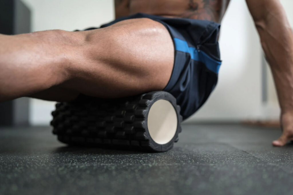 A person using a foam roller on their leg while sitting on the floor in a gym or workout space.