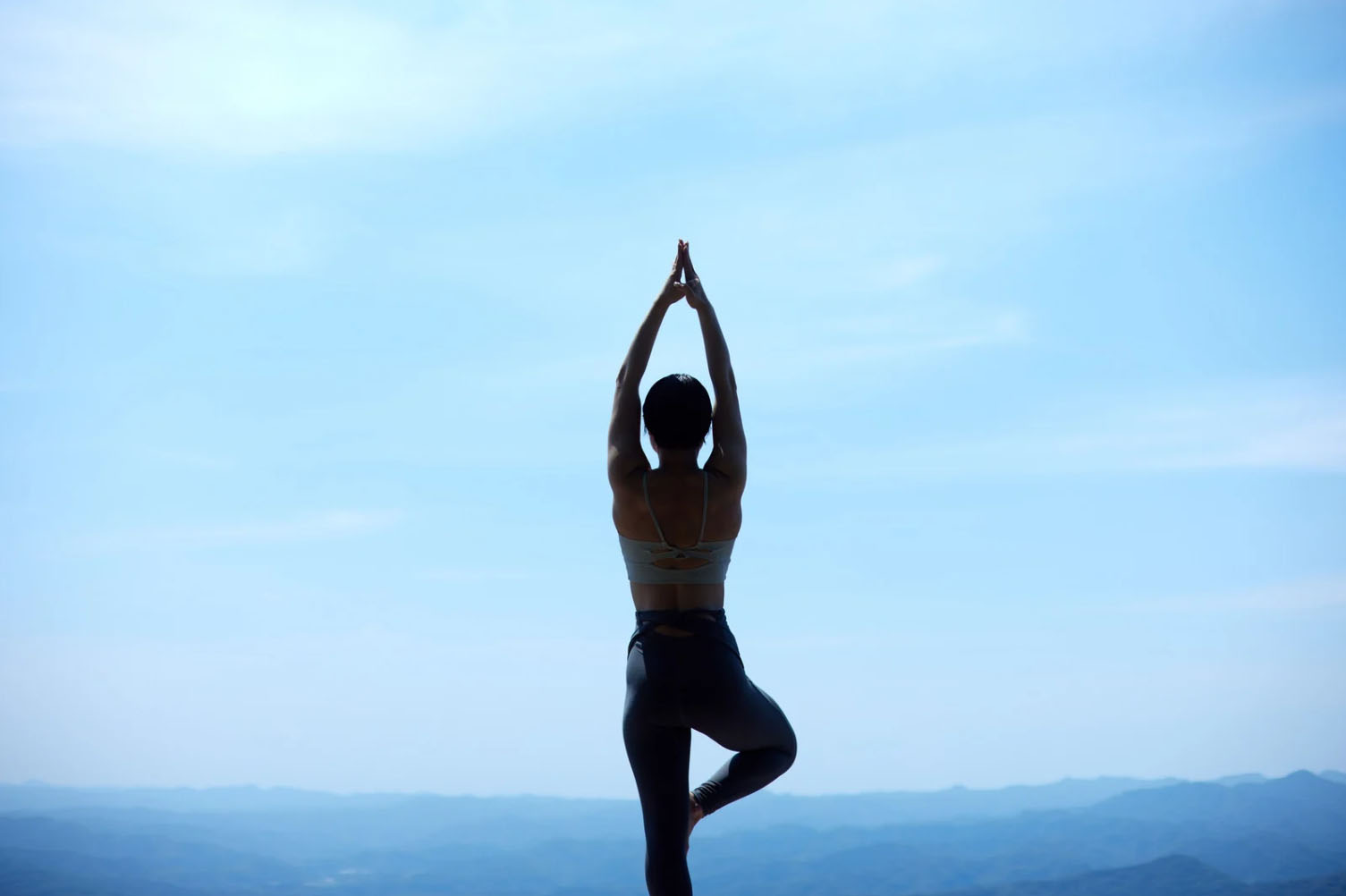 A person practicing yoga in a tree pose on a mountain top, with a clear blue sky and scenic landscape in the background.