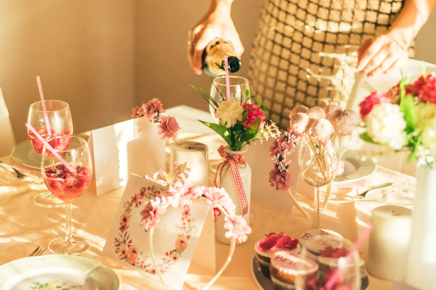 A beautifully set table with pink flowers, wine glasses, a bottle being poured, and decorative items including greeting cards and cupcakes, creating a festive and elegant atmosphere.