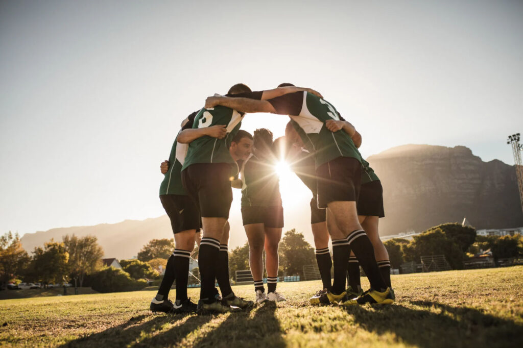 A group of six people, dressed in green sports uniforms, are standing in a huddle on a field with their hands together, against a backdrop of a sunset and mountains.