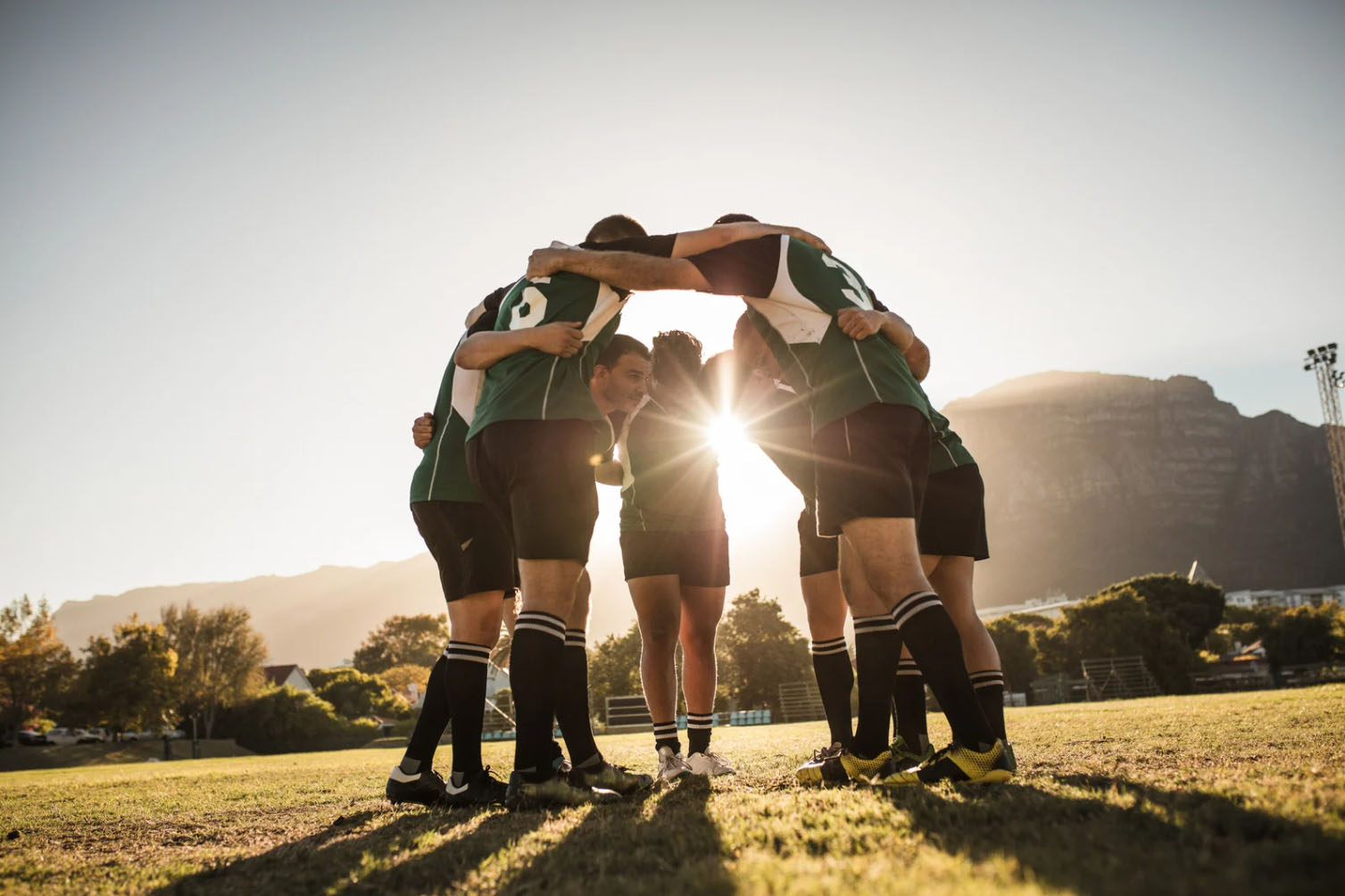 A group of six people, dressed in green sports uniforms, are standing in a huddle on a field with their hands together, against a backdrop of a sunset and mountains.