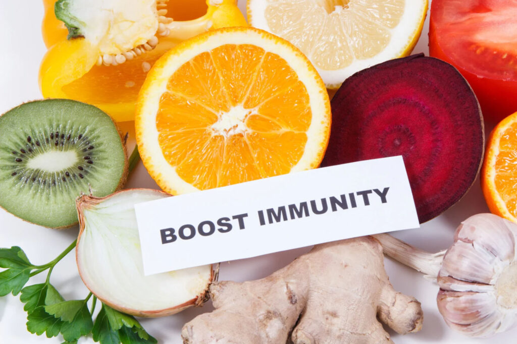A variety of colorful fruits and vegetables including orange, kiwi, lemon, bell pepper, garlic, onion, and beetroot arranged around a card with the text "BOOST IMMUNITY".