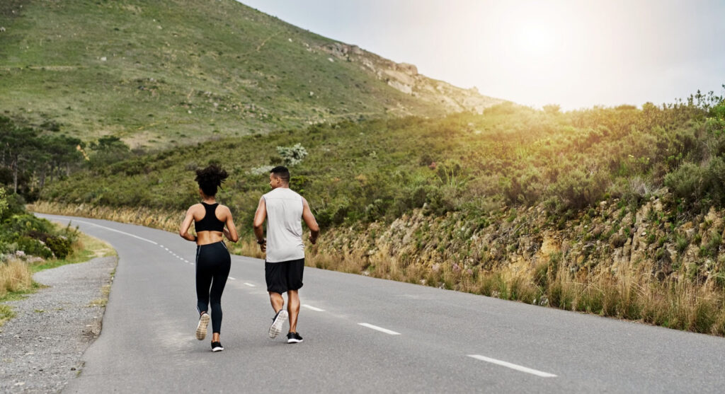 A man and woman jogging side by side on a scenic road surrounded by hills and greenery, with the sun shining in the background.