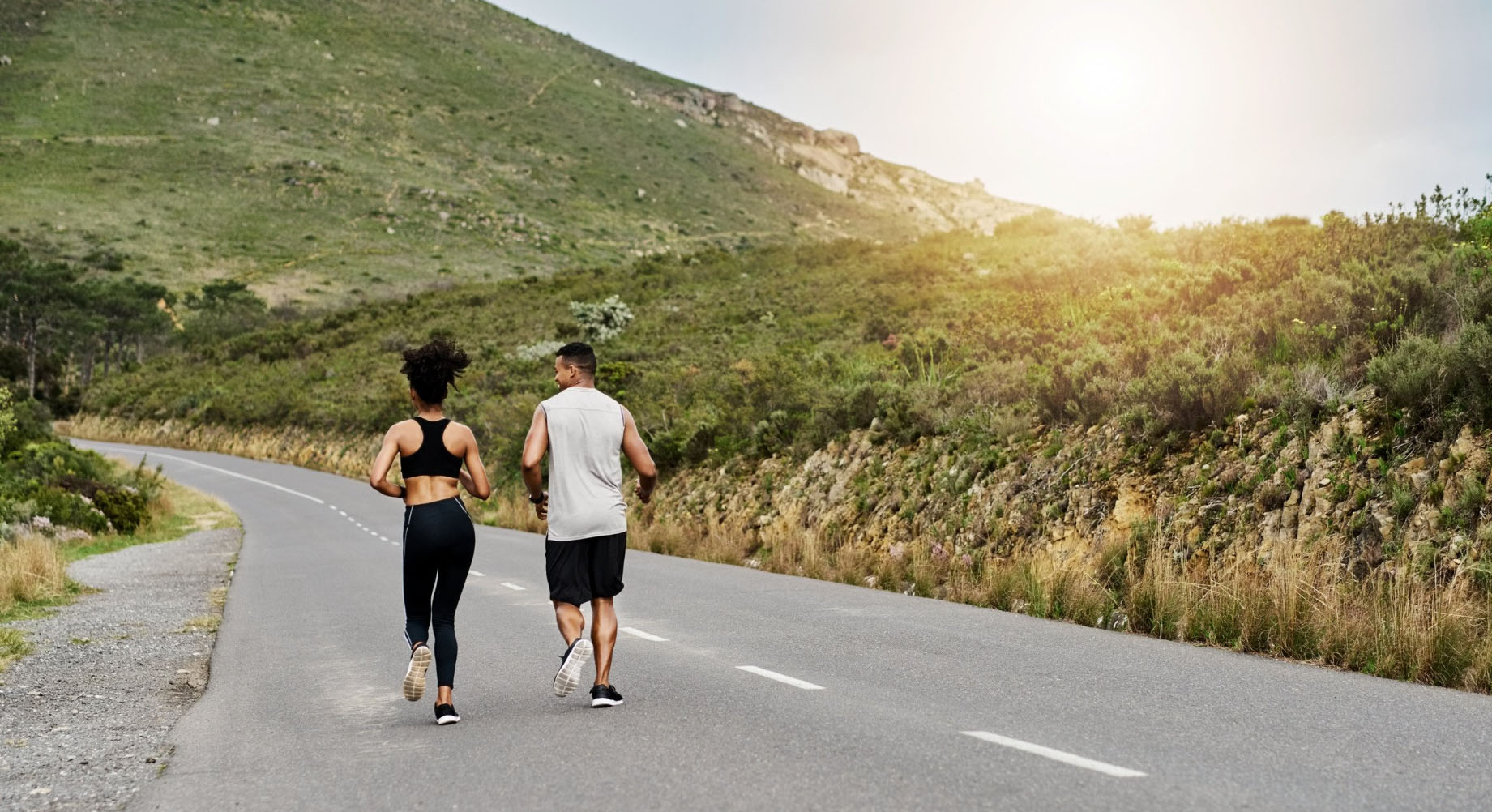 A man and woman jogging side by side on a scenic road surrounded by hills and greenery, with the sun shining in the background.