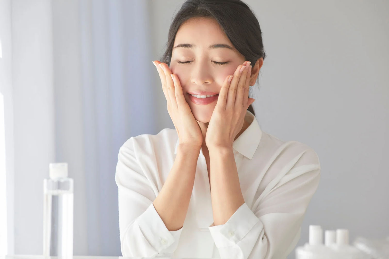 A woman with dark hair wearing a white shirt is gently pressing her hands on her face, smiling contently, with skincare products visible in the background.