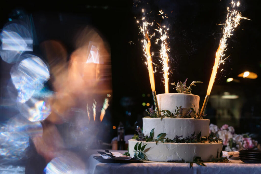 A three-tier white wedding cake decorated with greenery and flowers, topped with sparklers burning brightly in a dimly lit celebration setting.