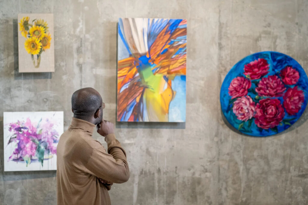 An individual is admiring colorful paintings displayed on a concrete wall, including floral artworks such as sunflowers, abstract patterns, and roses.