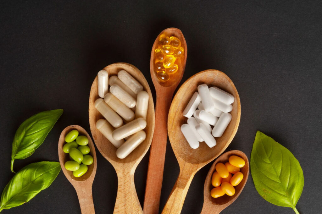 An assortment of dietary supplements in various shapes and colors placed on wooden spoons, with green leaves surrounding them, against a dark background.
