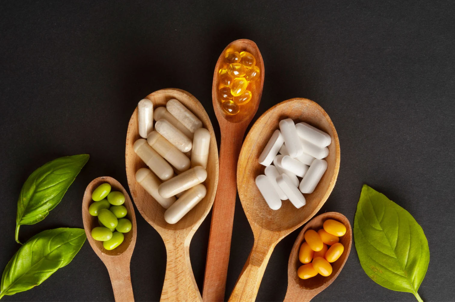 An assortment of dietary supplements in various shapes and colors placed on wooden spoons, with green leaves surrounding them, against a dark background.