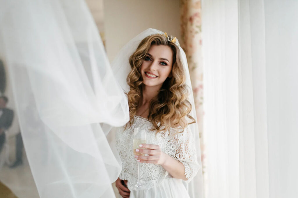A bride with wavy hair and a floral crown smiles while holding a glass of champagne. She is surrounded by flowing wedding veil fabric and a softly lit window, giving the scene a dreamy atmosphere.