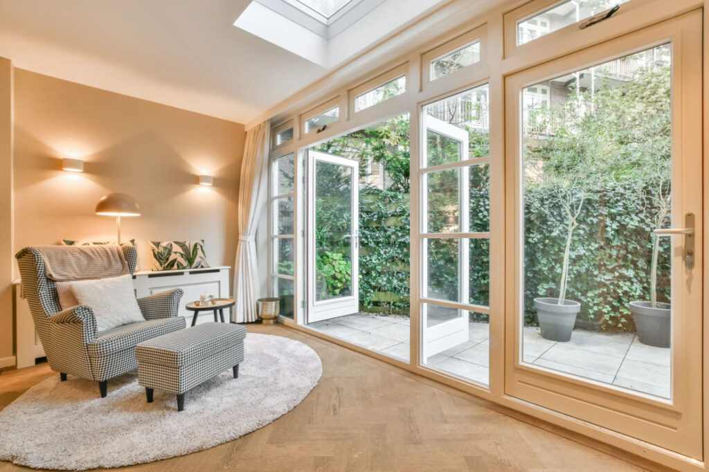 Bright and cozy living room with an armchair, ottoman, and floor lamp beside large glass doors opening to a garden patio.