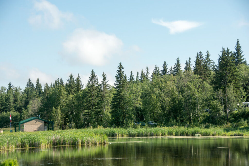 A serene landscape featuring a calm pond in the foreground, surrounded by lush greenery and tall trees. In the background, a small building with a Canadian flag is visible, partially obscured by the trees. The sky above is partly cloudy with clear blue patches.