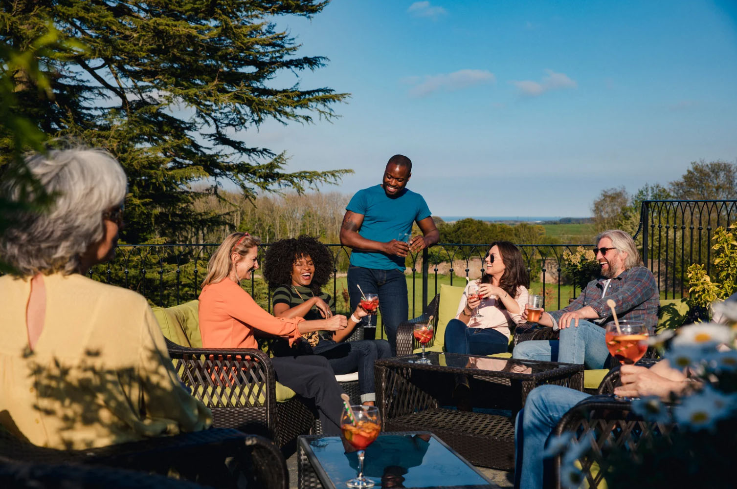 A group of six people, including both men and women, enjoy drinks and conversation outdoors on a sunny day. The group is seated on outdoor furniture, surrounded by greenery with a clear blue sky in the background.