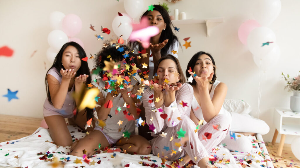Group of women in pajamas celebrating on a bed, blowing colorful confetti with balloons in the background.