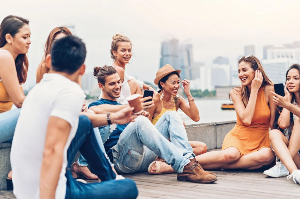 Group of young adults sitting outdoors near a waterfront, laughing and talking together while holding smartphones, with a city skyline in the background.