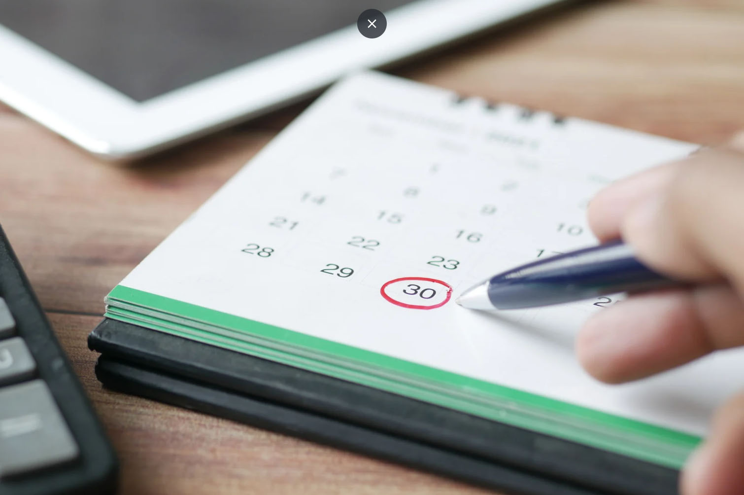 Close-up of a hand holding a pen and pointing at a calendar with the 30th day circled in red, with a tablet and calculator nearby on a desk.